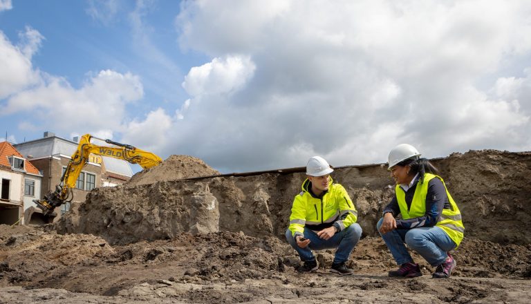 Twee Omgevingsdienst Haaglanden collega's op hun hurken bij een ontgrondingactiviteit. Daarbij staan ze op een bouwplaats met veel zand en staat er een afgraafkraan achter hun in beeld.