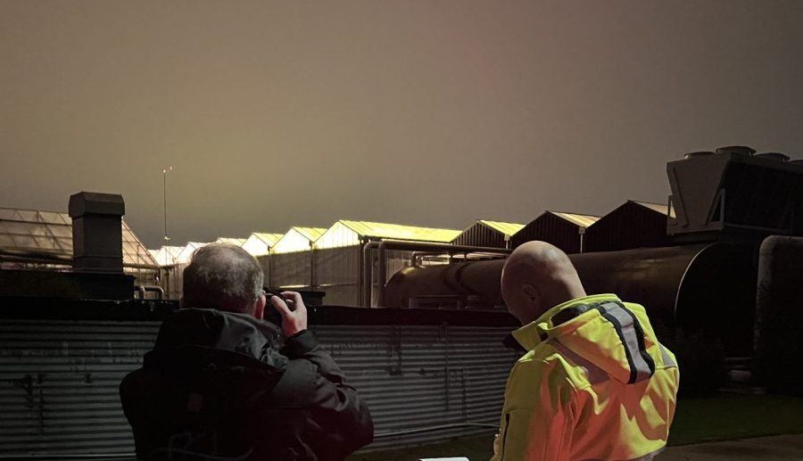 Twee toezichthouders tijdens een assimilatiebelichting controle in de nacht. De één noteert iets op papier en de ander maakt een foto van een broeikas dat licht uitstoot.